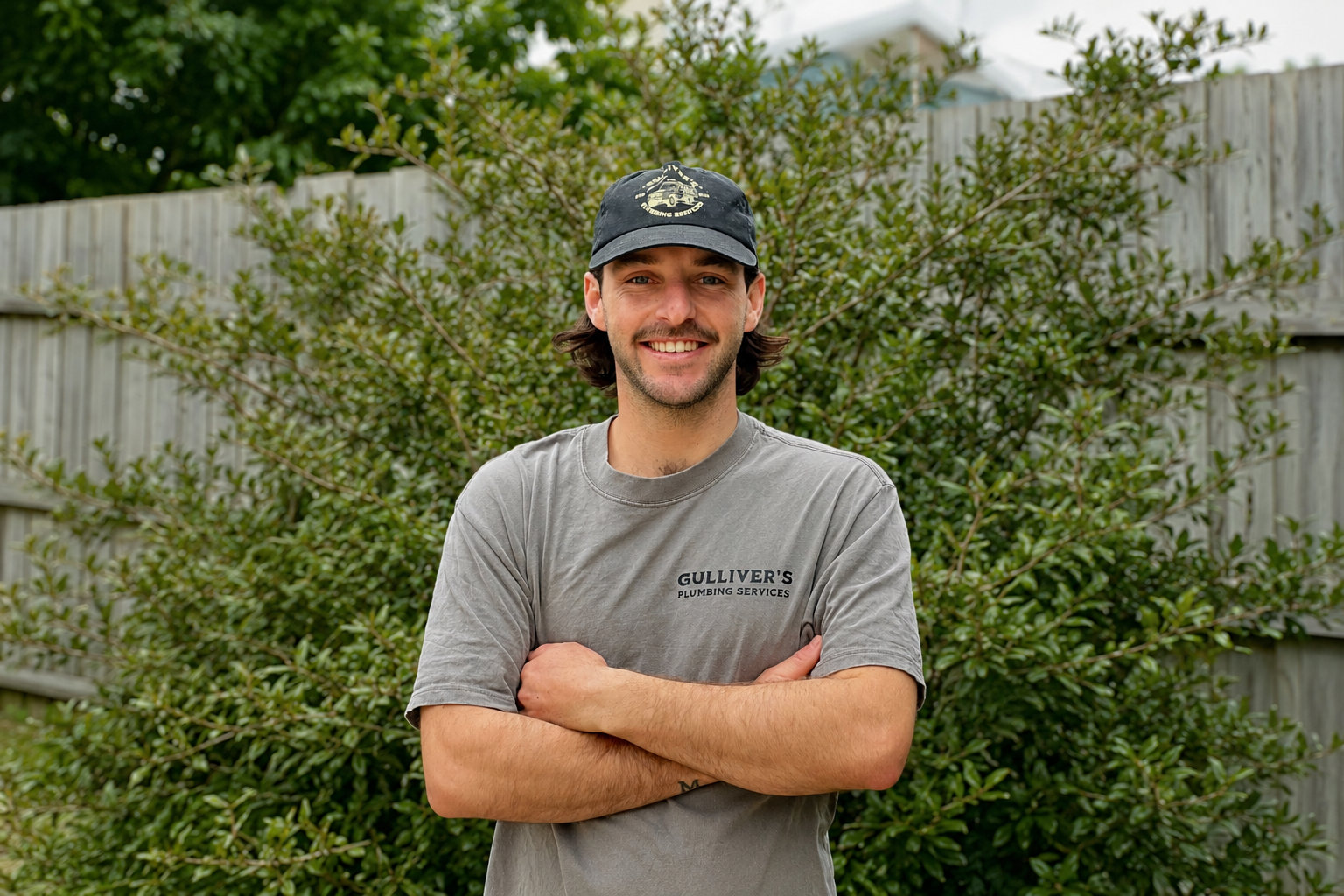 A man wearing a black cap and a grey “Gulliver’s Plumbing Services” shirt stands outside in front of a bush, smiling with arms crossed.